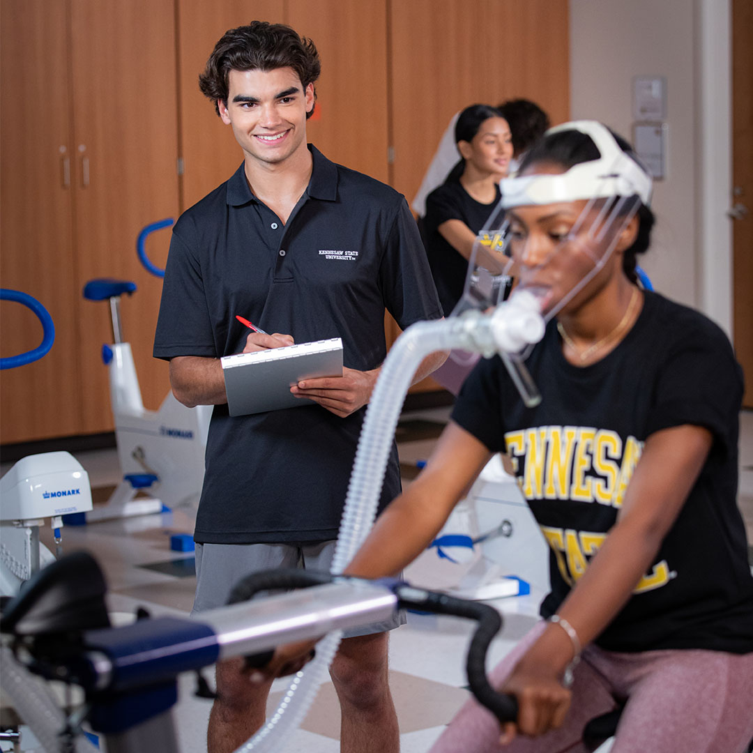 Faculty member monitoring biomechanics data on a tablet while a child runs through a motion capture system in a kinesiology research lab with sensors and analysis software displayed on a screen.