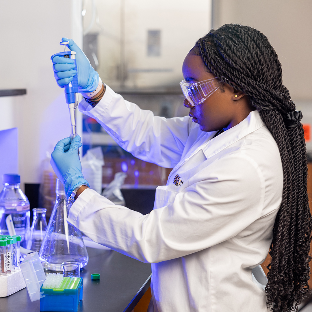 A laboratory researcher in a white lab coat uses a pipette to prepare samples at a workstation surrounded by beakers, test tubes, and scientific equipment.