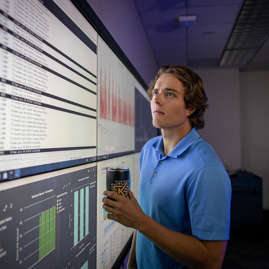 A man holding a cup is looking up at a bank of screens containing data and charts.