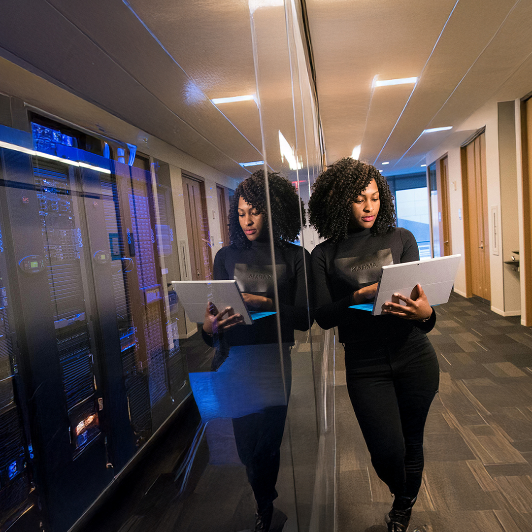 A woman holding a laptop standing in a hallway, leaning against a glassed-in server room.