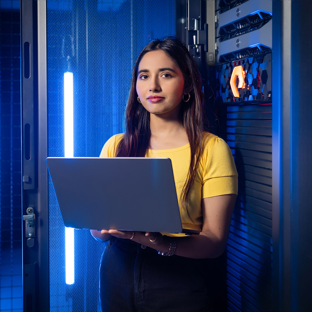 A woman stands in a server room, holding a laptop and looking focused on her work.