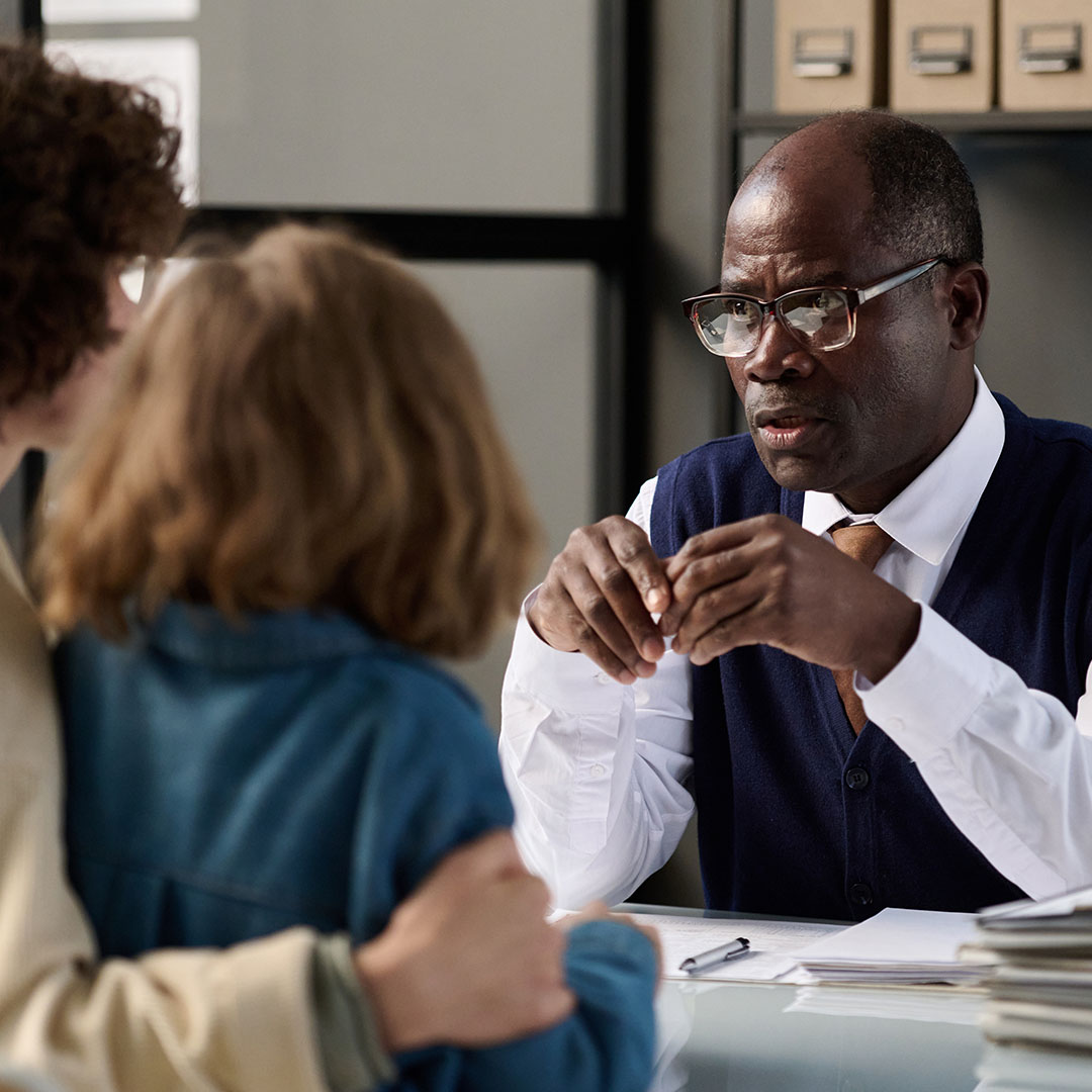 A professional seated at a desk speaking with two people across from the table, with paperwork and office storage visible in the background.