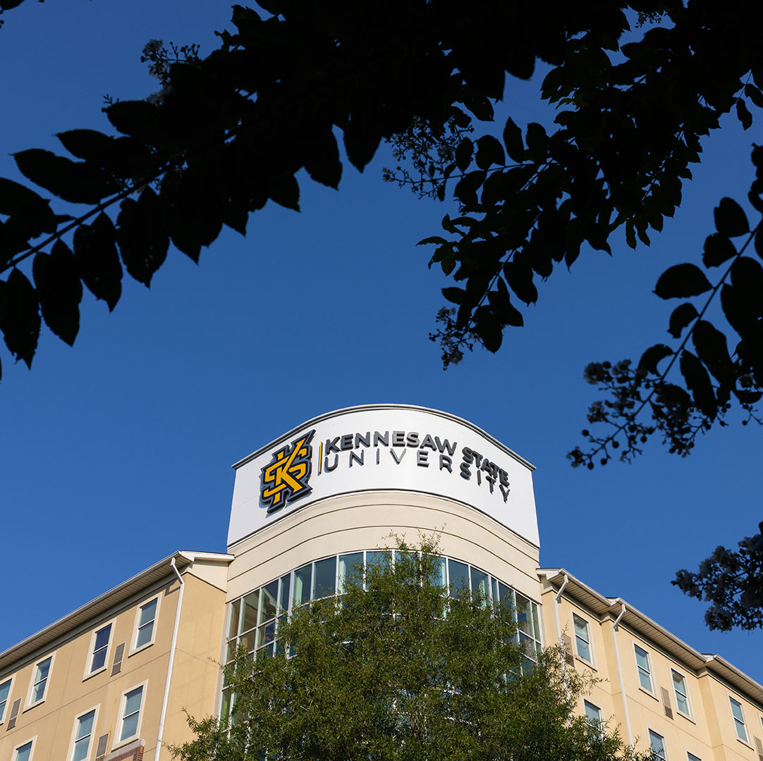Gold, black and white KSU building sign framed by tree limbs in dark blue sky.