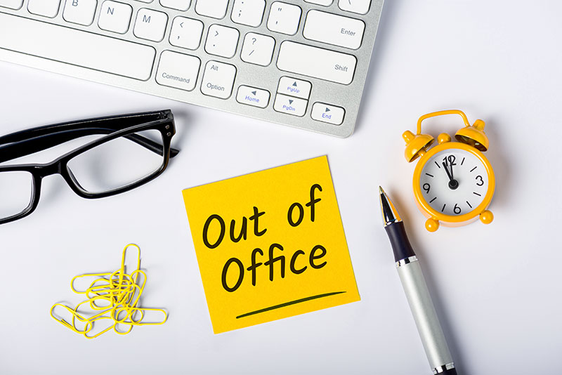 Office desk with a keyboard, glasses, paper clips, alarm clock, pen and a sticky note with out of office written on it.