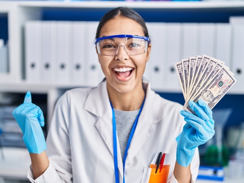 student scientist wearing a lab coat, goggles, and safety glasses holding $100 bills in one hand