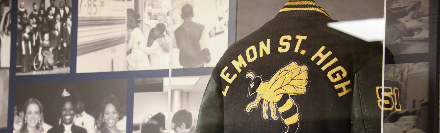 A museum display showing several black and white phots and a black and gold letterman jacket with Lemon St. High on the back.