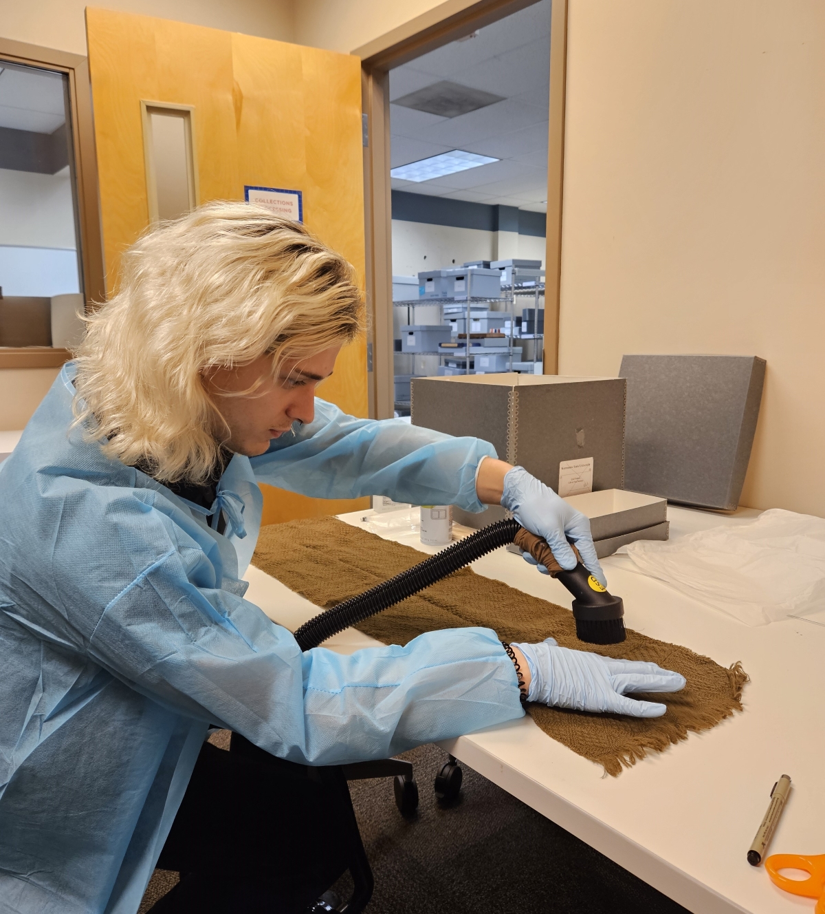 A person wearing protective gown and gloves uses a vacuum hose to clean a brown scarf 