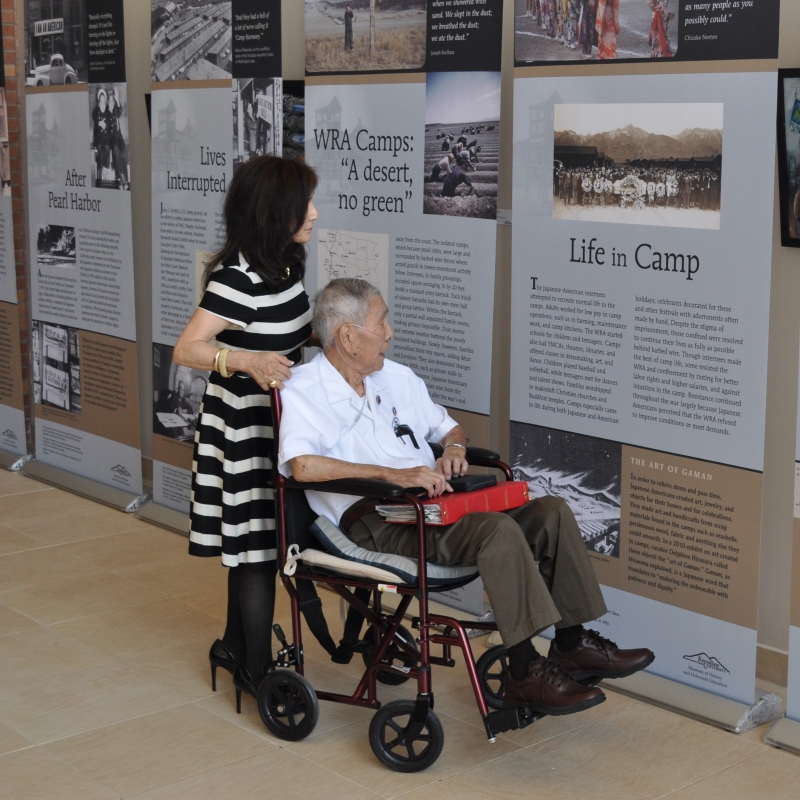 A woman pushes an old man in a wheelchair while they look at museum panels