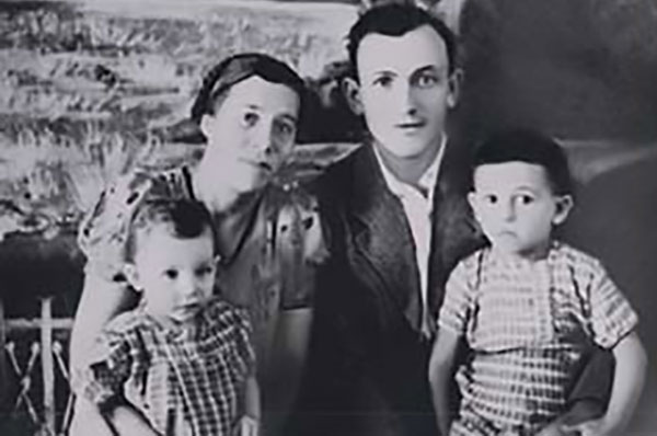 black and white photo of immigrant family of four seated in front of chalkboard with two young children on their laps.