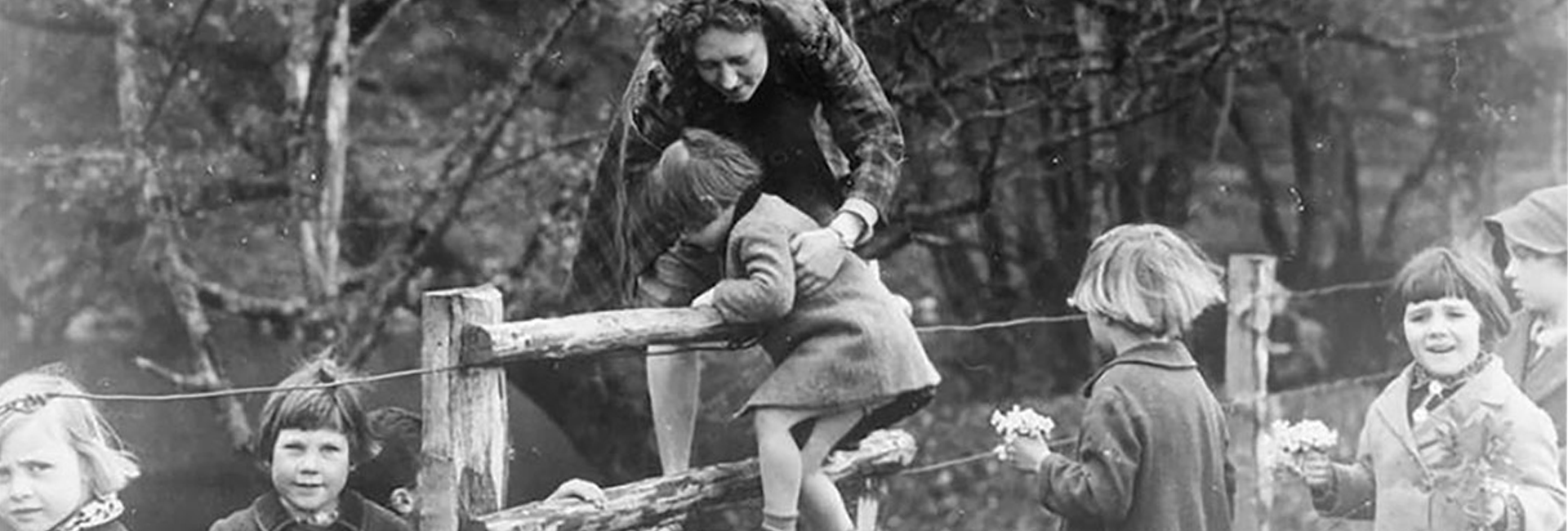 a black and white photo of a woman helping children cross over a fence