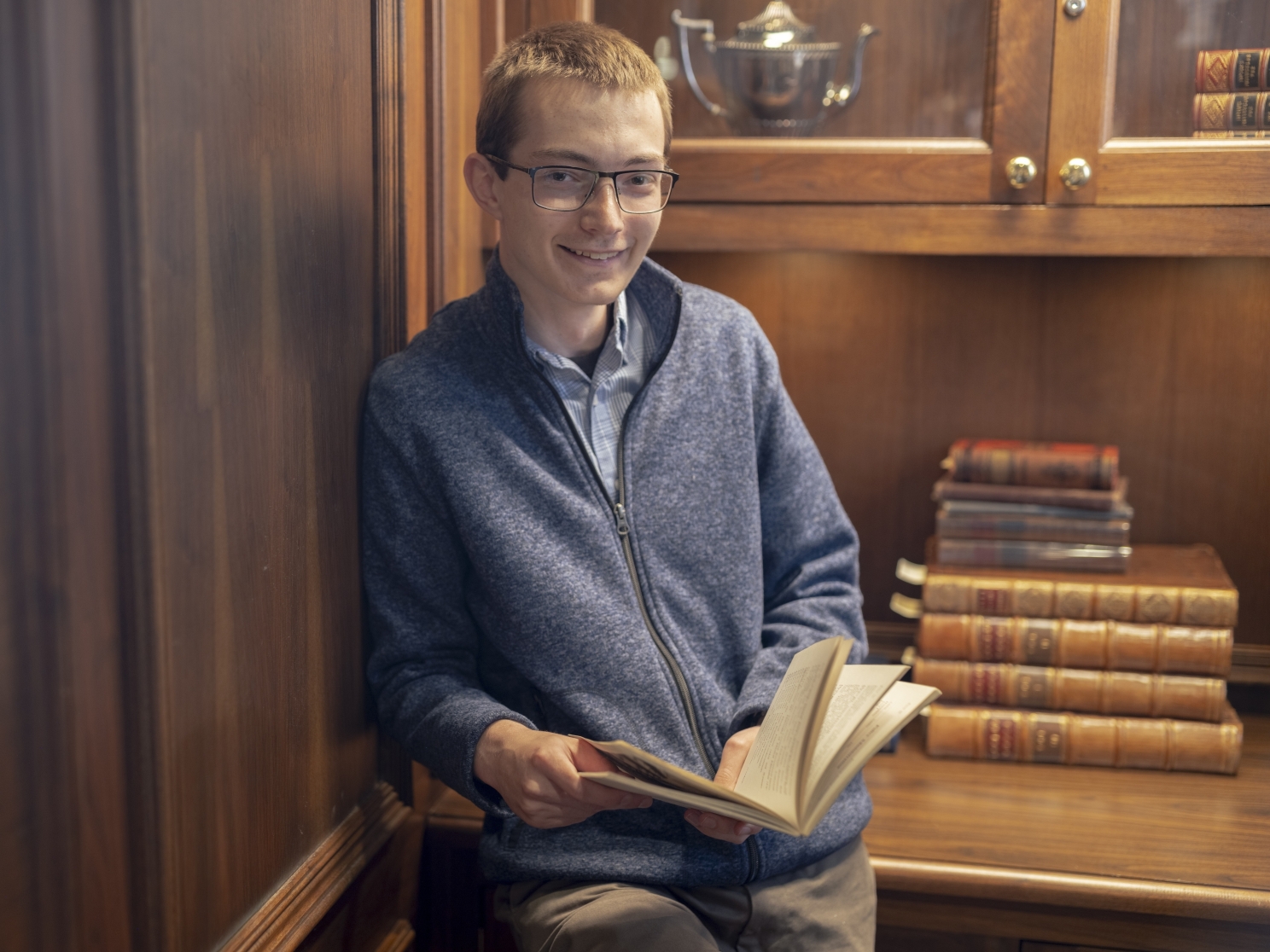 Young man standing in front of shelves of books while holding a book and smiling.