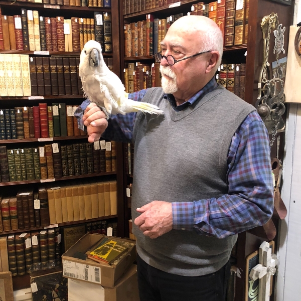 A large white parrot sits on the arm of an old man atanding in front od bookshelves.