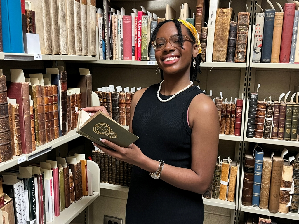 Young woman standing in front of shelves of books while holding a book and smiling.