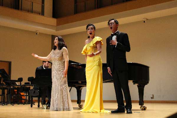 Three people in formal wear singing in a concert hall in front of a piano.