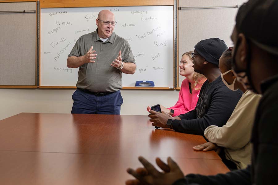 Photo of Neil Duchac standing classroom with students sitting around table