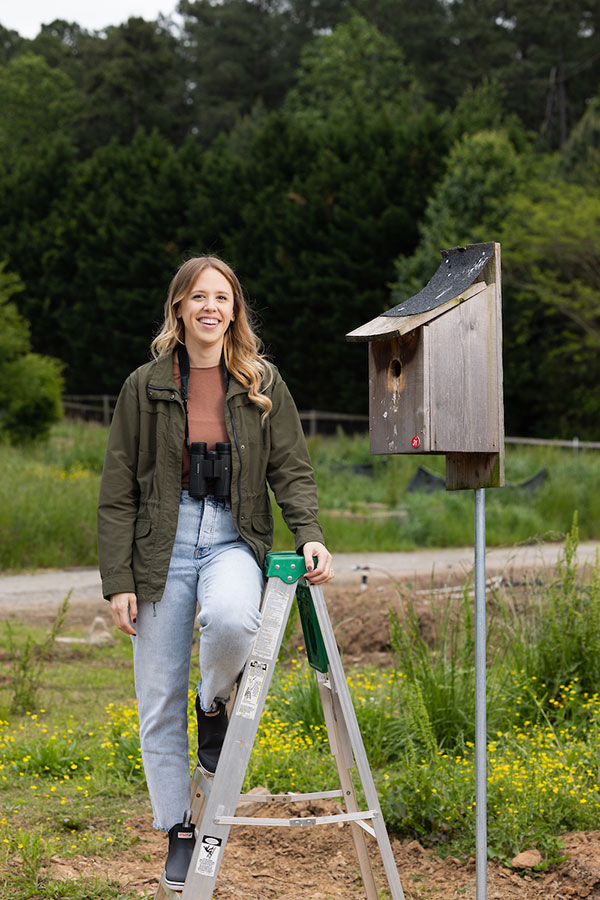 Photo of Rachel Kaplan standing on ladder beside a birdhouse at the KSU Field State