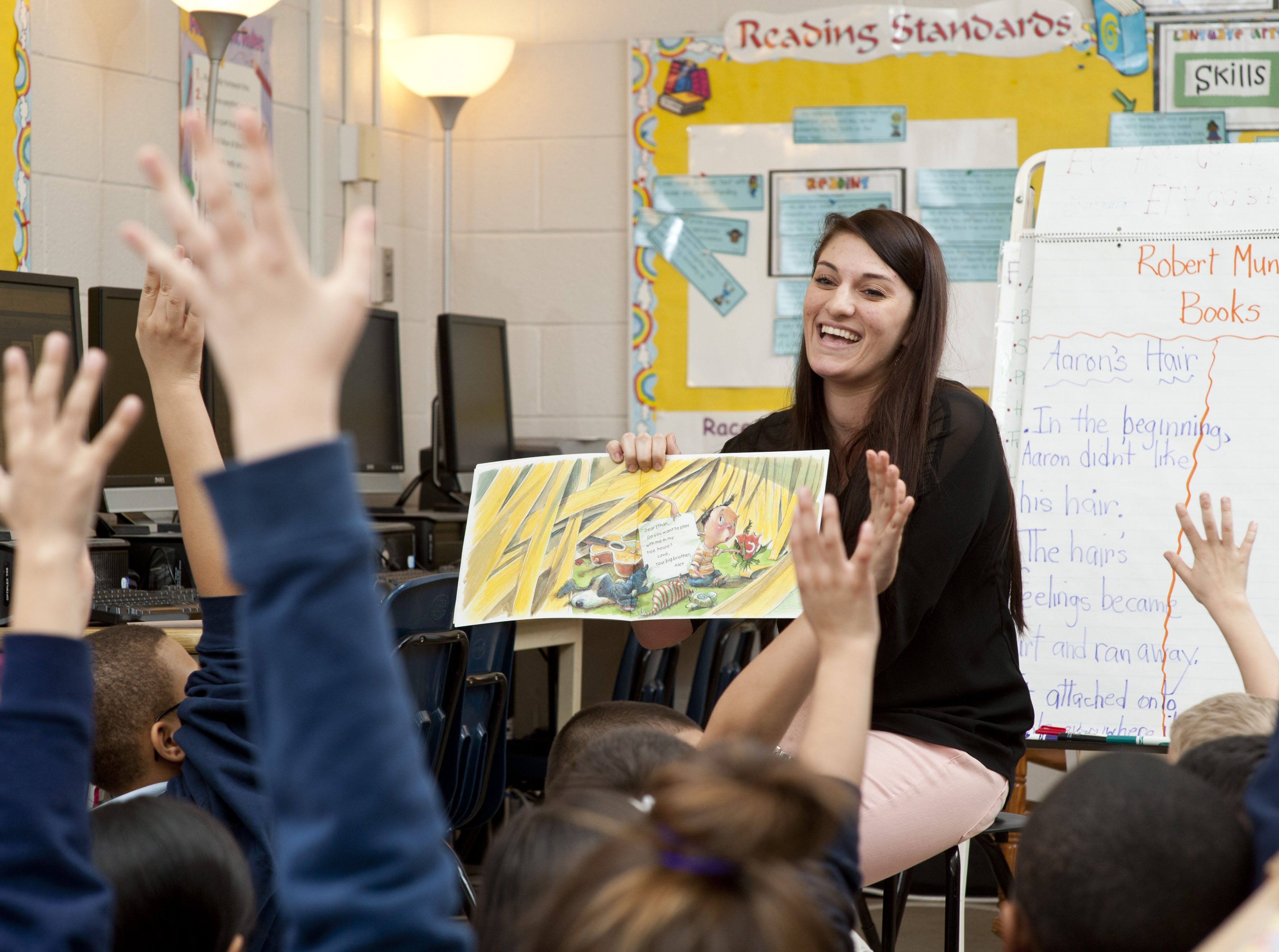 female teacher reading colorful book as she smiles to students with arms raised