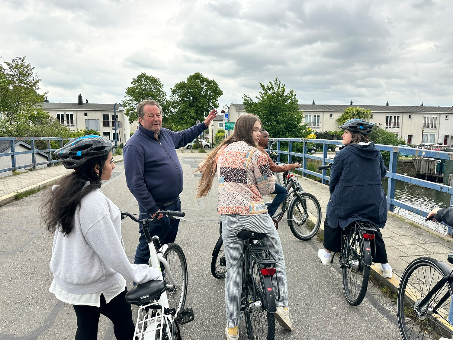 A group of cyclists pause on a bridge under overcast skies. A man gestures ahead, leading the group. The mood is relaxed and communal.