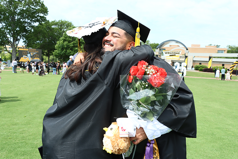 KSU graduate wearing a cap and gown hugs another person on a sunny lawn, holding a bouquet of red roses and a teddy bear, conveying joy and celebration.