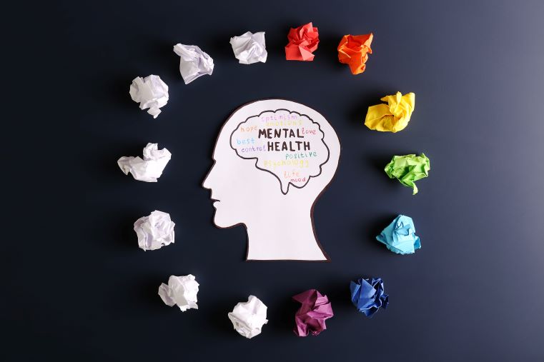 A paper cutout of a human head with "Mental Health" and related words inside the brain, surrounded by crumpled paper on a dark background.