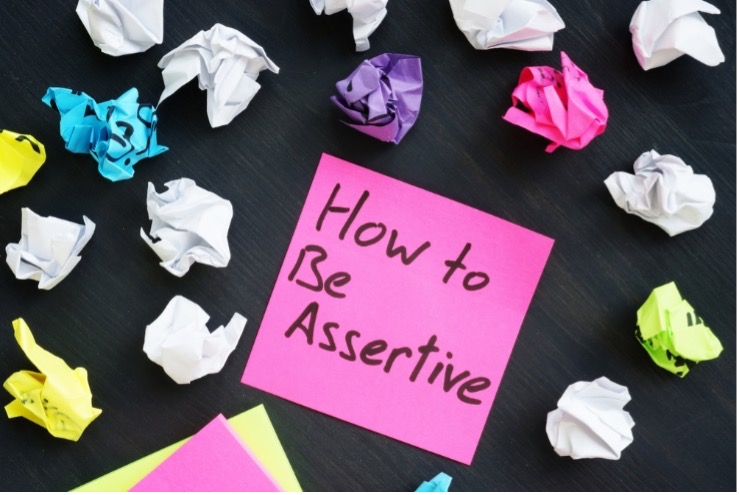 A pink sticky note with the words "How to Be Assertive" surrounded by colorful balled up pieces of paper.