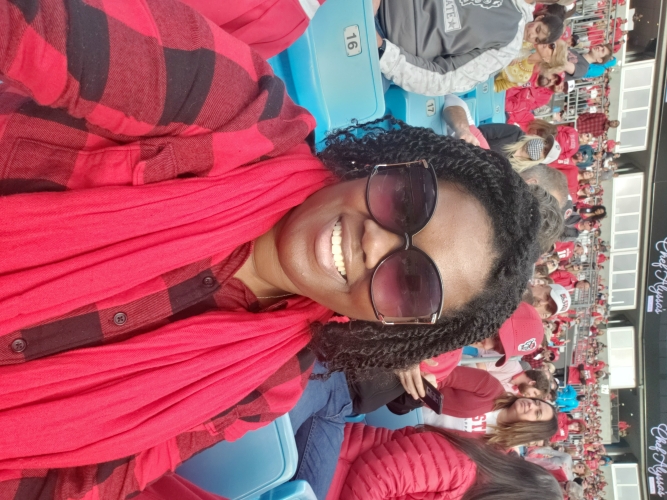 Alicia McGee wearing a red scarf and red-and-black plaid shirt, seated in a stadium with light blue seats. The background shows a crowd of people dressed mostly in red.