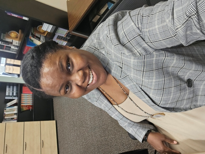 Alicia McGee dressed in a gray plaid blazer over a beige top, sitting at a desk in an office environment.