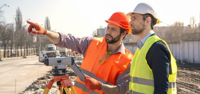 Two male land surveyors with a device working on a construction site in a helmet.