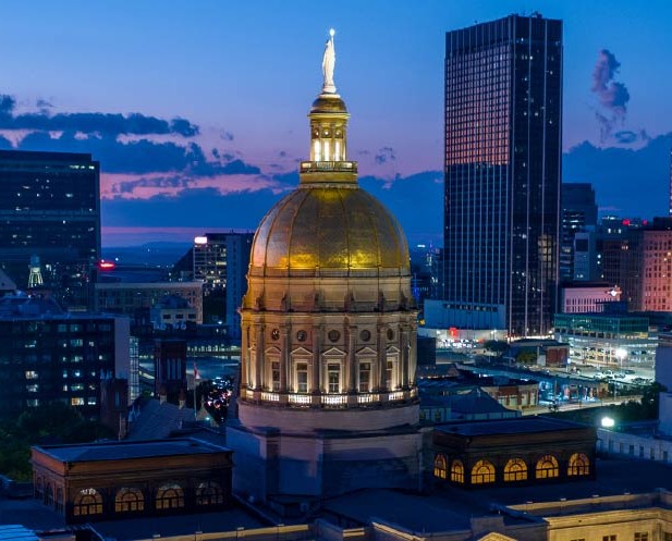 Georgia State Capitol building with its gold dome illuminated at dusk, surrounded by city skyscrapers and a colorful evening sky.