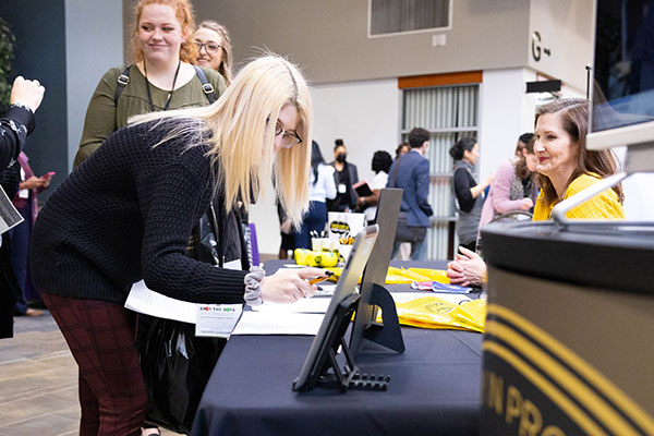KSU student leaning over a table at an event, writing on a form near a monitor and informational materials, with other attendees visible in the background.