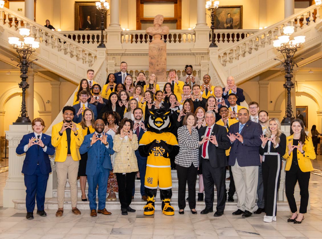 Kennesaw State administration and students standing on the main marble steps inside the Georgia capitol building making owl eyes with their hands. Srappy Owl mascot stands at the very front
