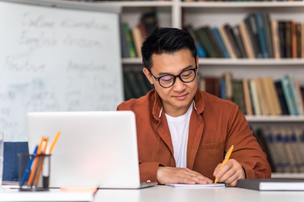 Teacher writing and using laptop in a classroom setting.