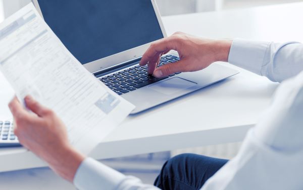 Man typing on a laptop and holding a form in his hand