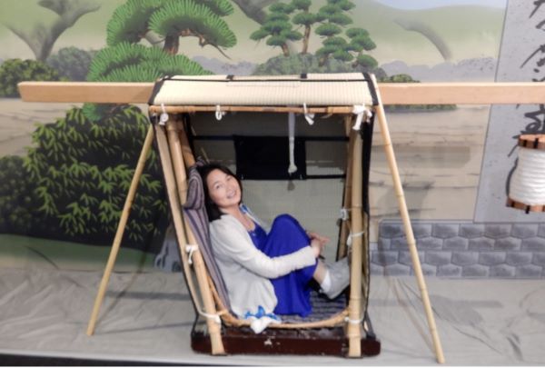 Woman sitting inside a traditional Japanese palanquin (kago) with a painted backdrop.