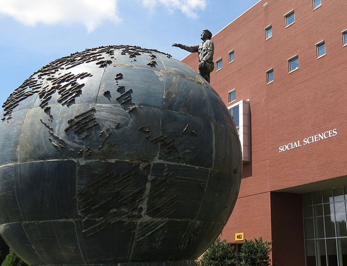A large bronze globe sculpture stands in front of KSU Social Sciences building and a statue of a man is positioned on top of the globe, reaching out with one arm