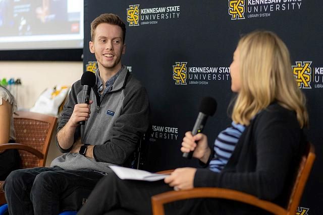 Two people holding microphones sitting in front of a black background with the KSU logo