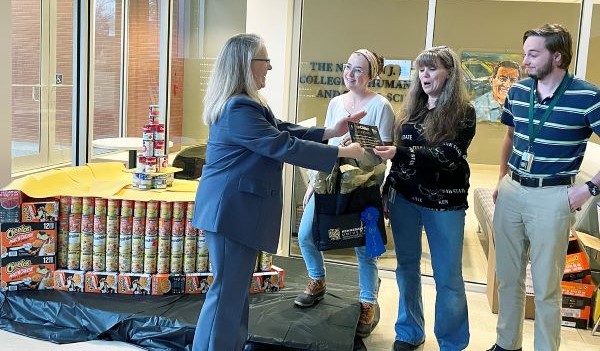A woman in a blue suit hands an item to a group of three people near a display of canned goods.