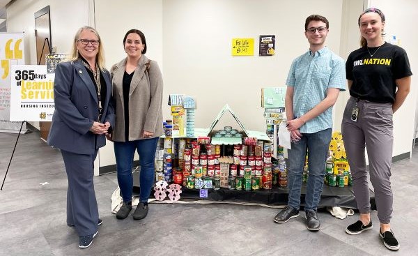 Four people stand in front of a pyramid made of canned goods and other food items in a hallway.
