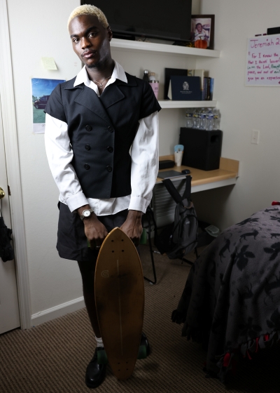Amorie Calixte holds a longboard skateboard vertically by its trucks in a room with shelves containing water bottles, books, and personal items. 