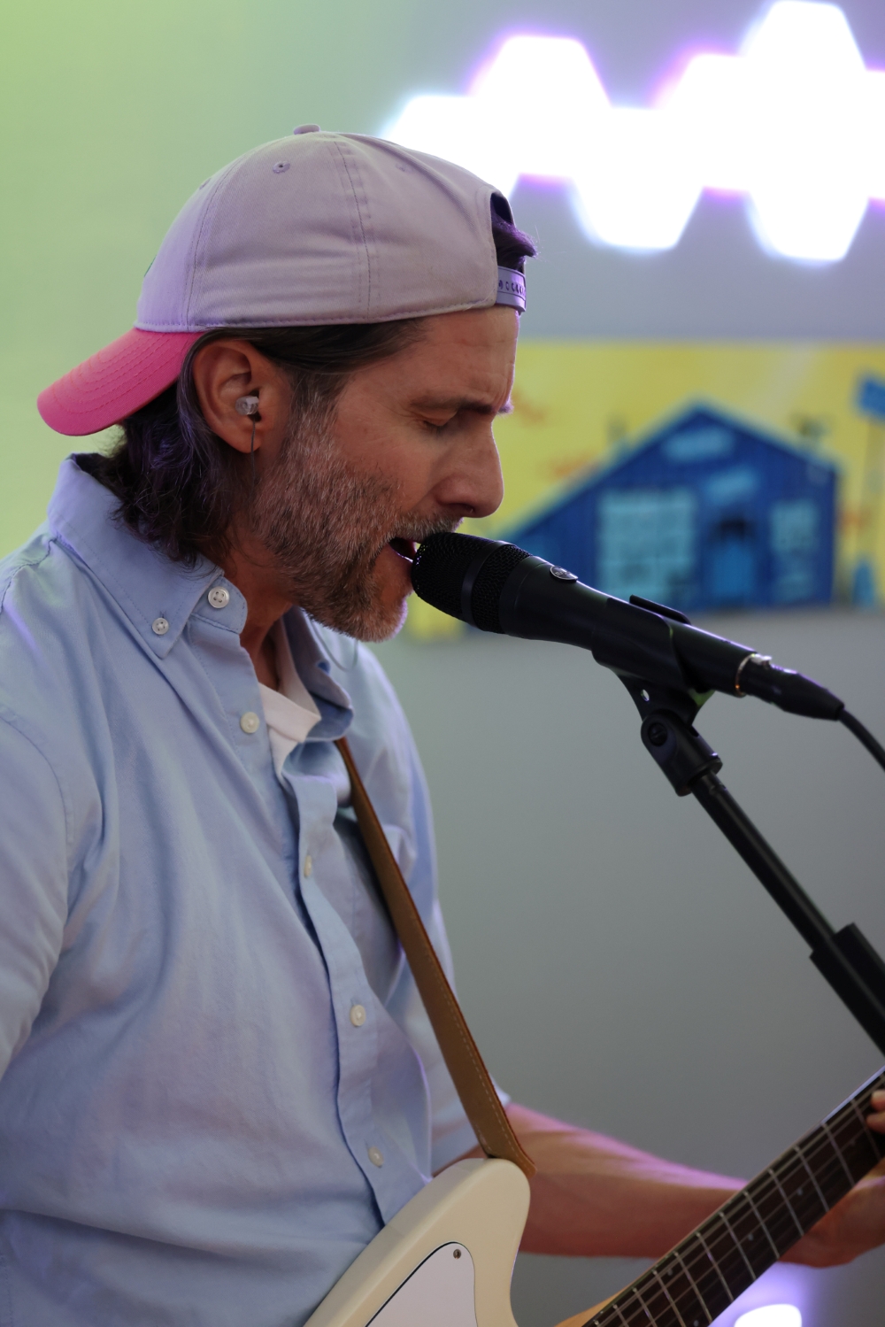 Kenneth White wearing a light blue button-up shirt and a backward pink and gray cap, singing into a microphone while playing an electric guitar. The background features a colorful wall with geometric shapes.