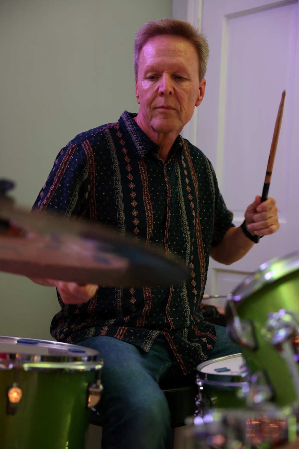 Kerwin Swint in a patterned shirt playing a green drum set, holding drumsticks in both hands