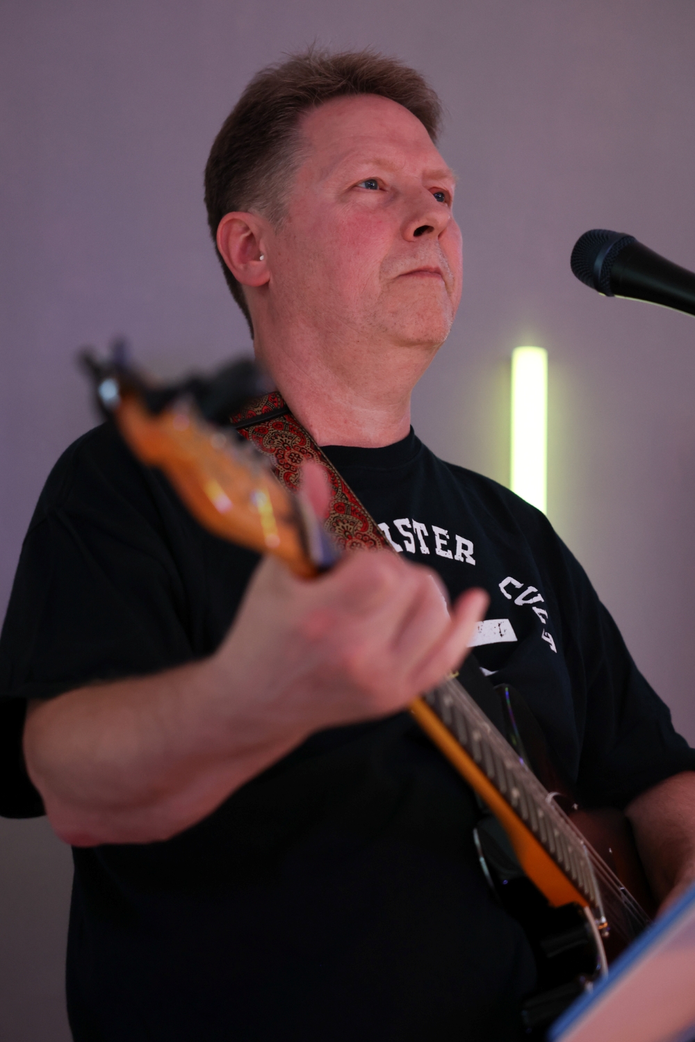 Tom Rotnem wearing a black t-shirt with text, playing an electric guitar. One hand is on the guitar neck and the other is strumming, with a focused expression.