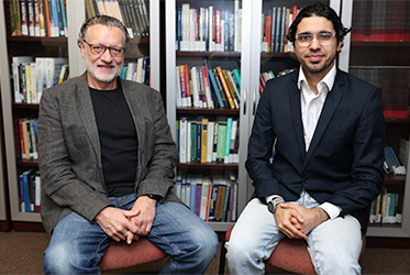 Professor Volker Franke and Amer Alnajar seated side by side in front of bookshelves filled with books. Franke on the left is wearing a dark blazer over a black shirt with jeans, and Alnajar on the right is wearing a dark suit jacket over a white shirt with light-colored pants. Both are sitting on chairs with their hands resting on their laps.