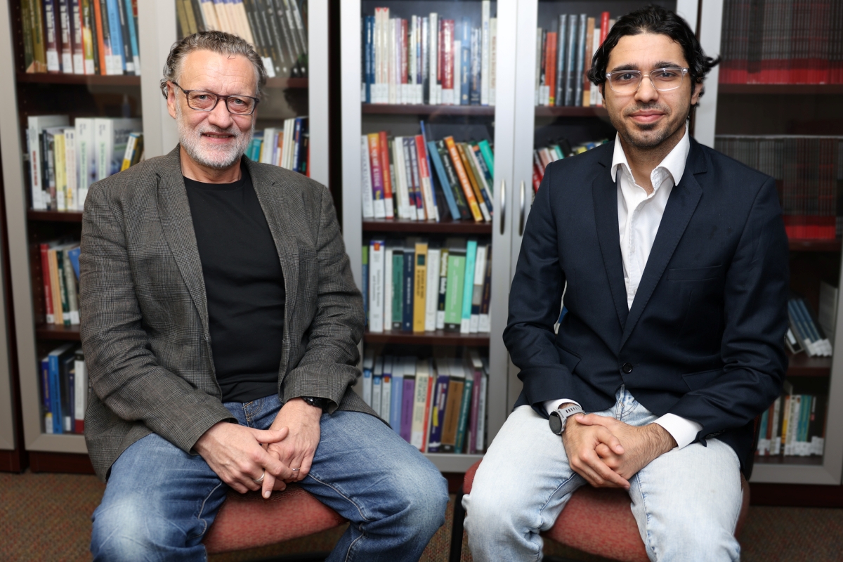 Professor Volker Franke and Amer Alnajar seated side by side in front of bookshelves filled with books. Franke on the left is wearing a dark blazer over a black shirt with jeans, and Alnajar on the right is wearing a dark suit jacket over a white shirt with light-colored pants. Both are sitting on chairs with their hands resting on their laps.