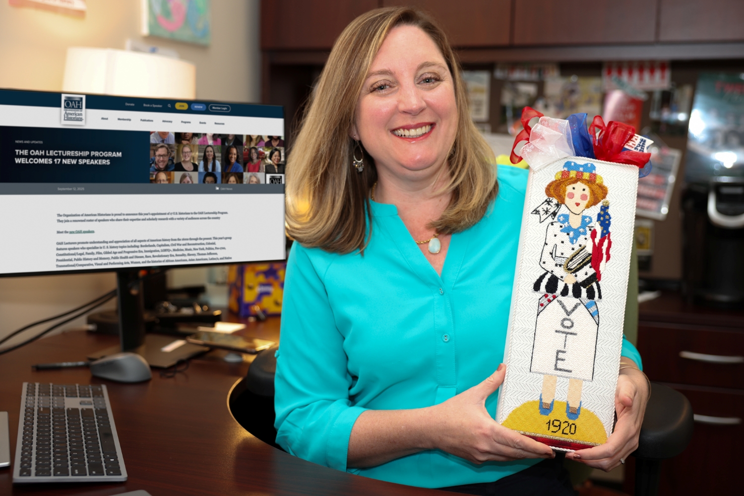 Lauren MacIvor Thompson wearing a teal blouse is seated at a desk holding a rectangular decorative item featuring an illustration of a figure in a white dress with the word “VOTE” and the year “1920” on it, adorned with red, white, and blue ribbons. A computer monitor on the desk displays a webpage titled “The OAH Lectureship Program Welcomes 7 New Speakers.” Office shelves and various items are visible in the background.