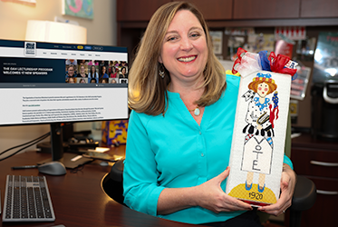 Lauren MacIvor Thompson wearing a teal blouse is seated at a desk holding a rectangular decorative item featuring an illustration of a figure in a white dress with the word “VOTE” and the year “1920” on it, adorned with red, white, and blue ribbons. A computer monitor on the desk displays a webpage titled “The OAH Lectureship Program Welcomes 7 New Speakers.” Office shelves and various items are visible in the background.