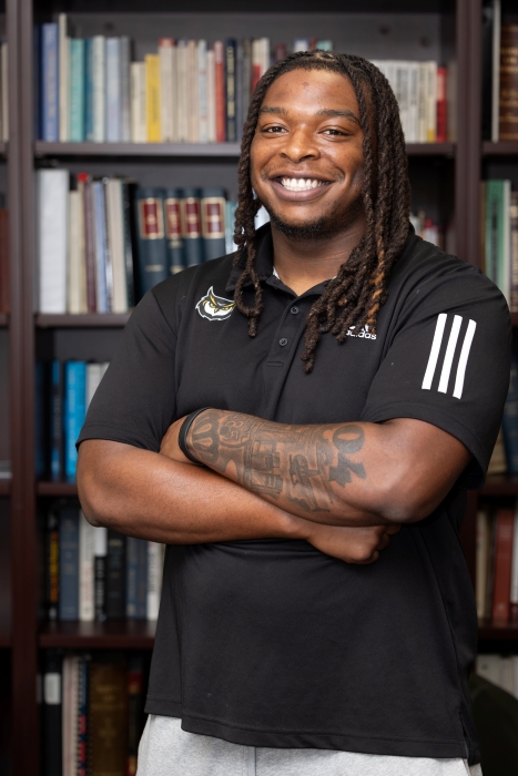 Tylon Dunlap in black KSU polo with owl logo and tattooed forearm stands with arms crossed in front of a bookshelf.