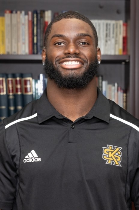 Coleman Bennett in black KSU polo with KS logo and Adidas branding stands in front of a bookshelf filled with books