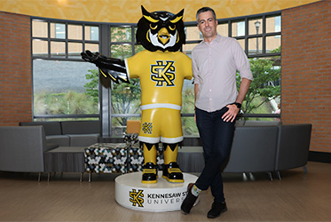 Marcelo Medeiros standing next to a large Kennesaw State University Scrappy Owl statue inside a modern lounge area with brick walls, large windows, and seating in the background.
