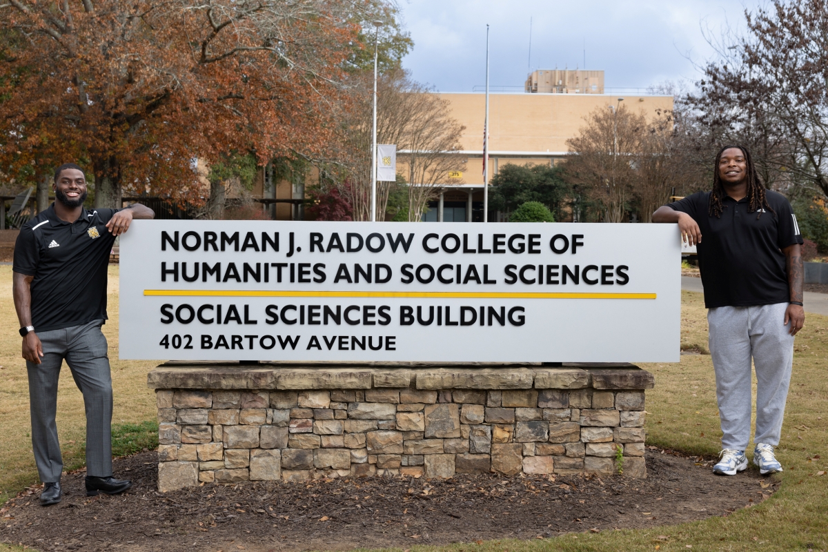 Two Kennesaw State University football players Coleman Bennett and Tylon Dunlap stand outdoors on either side of a stone sign that reads ‘Norman J. Radow College of Humanities and Social Sciences, Social Sciences Building, 402 Bartow Avenue.’ Trees with autumn leaves and a campus building are visible in the background